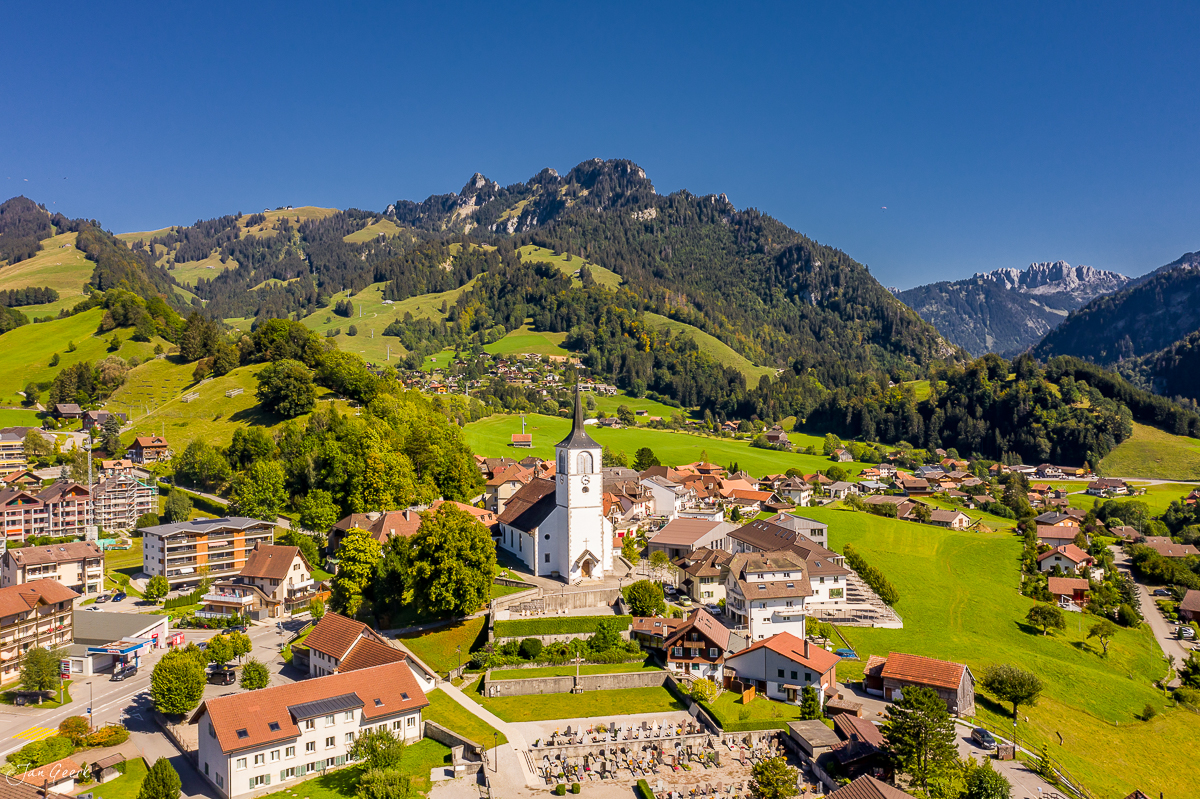 Luftaufnahme des Dorfes Charmey mit Kirche, grünen Alpenwiesen und Bergen im Sommer in der Westschweiz – Jan Geerk Fotograf Basel