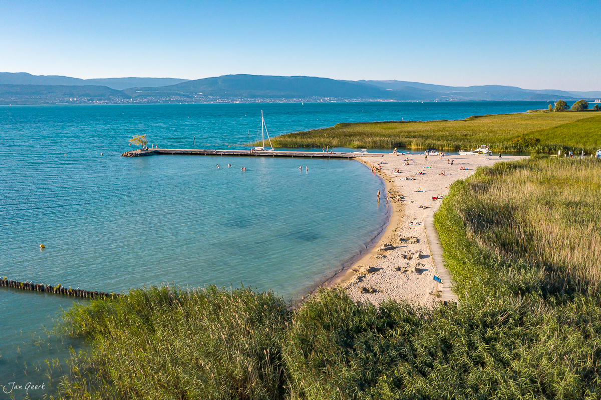 Sandstrand mit Badegästen am Neuenburgersee in Estavayer-le-Lac, türkisblaues Wasser, grüne Wiesen, Segelboot, Sommerlicht, Westschweiz, Schweiz – Jan Geerk Fotograf Basel