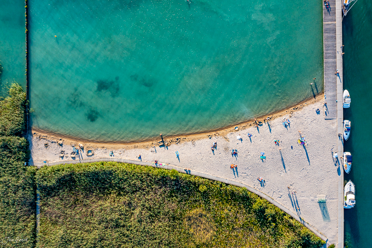 Luftaufnahme Badestrand Neuenburgersee mit Booten, türkisblauem Wasser und grüner Vegetation, Sommer, Westschweiz – Jan Geerk Fotograf Basel