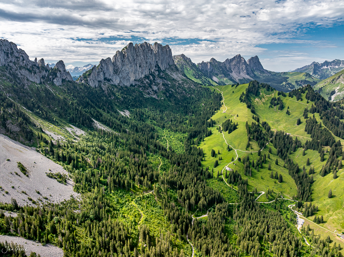 Luftaufnahme der Gastlosen mit grünen Alpweiden, Fichten und Serpentinenwegen, Sommerlicht, Prealpen Schweiz – Jan Geerk Fotograf Basel