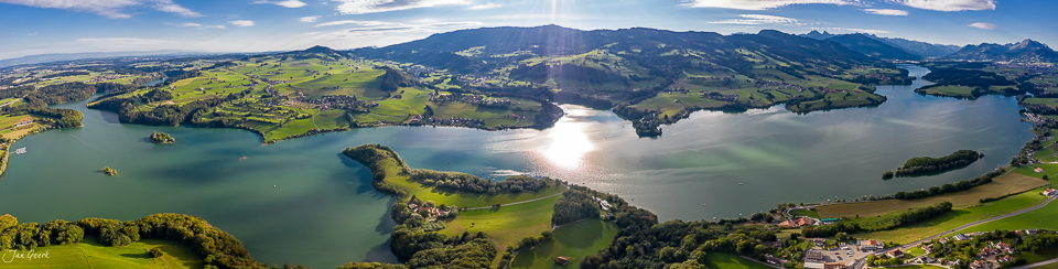 Luftaufnahme des Greyerzersees mit grünen Uferlandschaften, Inseln und Bergen in goldenem Kantlicht, Westschweiz – Jan Geerk Fotograf Basel