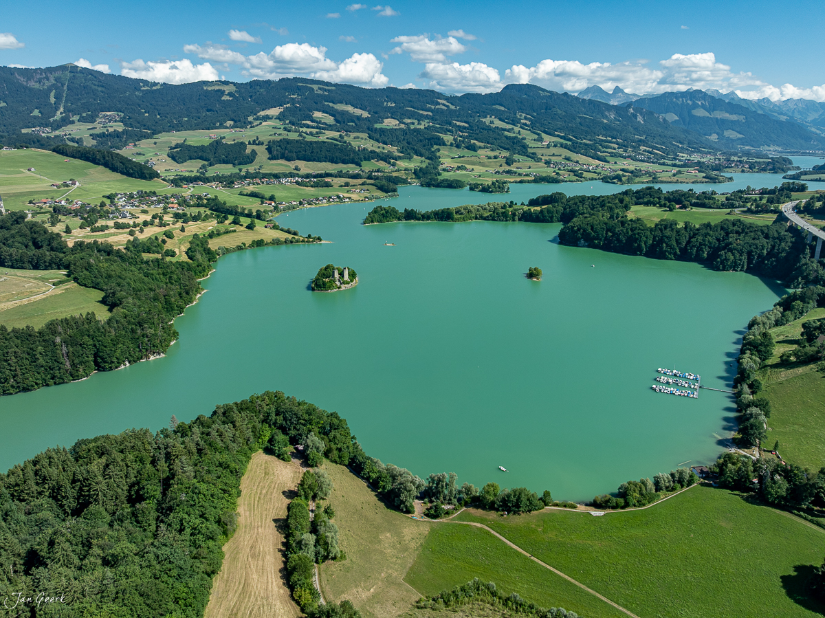 Luftaufnahme des Greyerzersees mit türkisfarbenem Wasser, umgeben von grünen Wiesen und Wäldern, Bergpanorama im Hintergrund, Sommeraufnahme Schweiz – Jan Geerk Fotograf Basel
