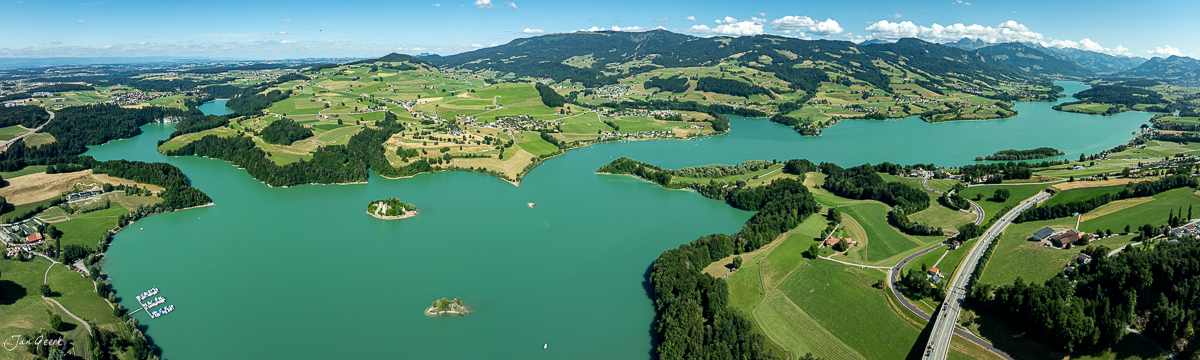 Luftaufnahme Greyerzersee mit Inseln, grünen Uferlandschaften und Bergen, Sommerstimmung, Freiburg Schweiz – Jan Geerk Fotograf Basel