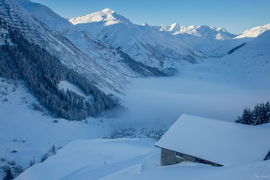 Verschneite Berglandschaft mit Nebel in Andermatt, Schweiz. Winterliche Alpenszenerie mit schneebedeckten Hängen und Talhäusern – Jan Geerk Fotograf Basel