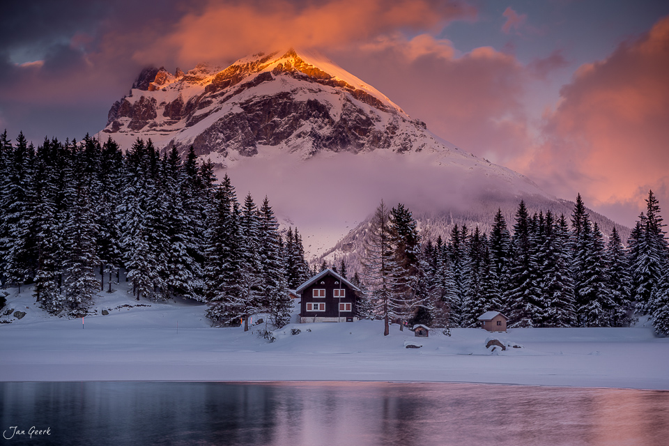 Schneebedeckter Bergsee mit rotem Holzhaus, verschneiter Tanne und schneebedecktem Berg bei Sonnenaufgang am Arnisee in der Zentralschweiz, Schweiz – Jan Geerk Fotograf Basel