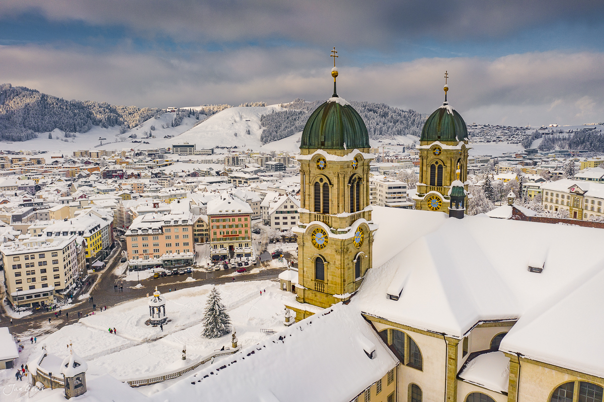 Kloster Einsiedeln mit schneebedecktem Dach und gelben Zwiebeltürmen, Luftaufnahme im Winter, Schweiz – Jan Geerk Fotograf Basel