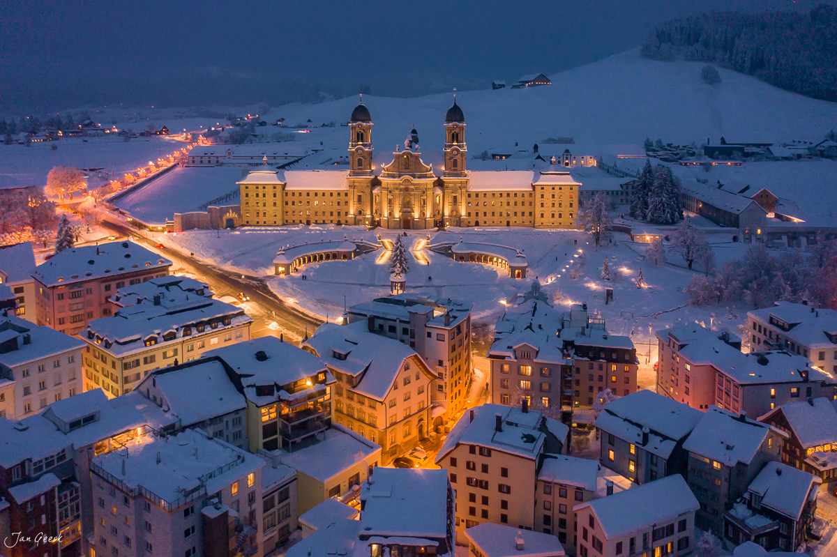 Kloster Einsiedeln in der Winterdämmerung mit schneebedecktem Tal und beleuchteten Gebäuden – Luftaufnahme Schweiz Jan Geerk Fotograf Basel