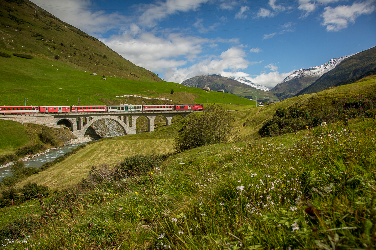 Roter Zug auf Steinviadukt über Bach, grüne Berglandschaft mit Schneebergen beim Oberalppass, Schweiz – Jan Geerk Fotograf Basel