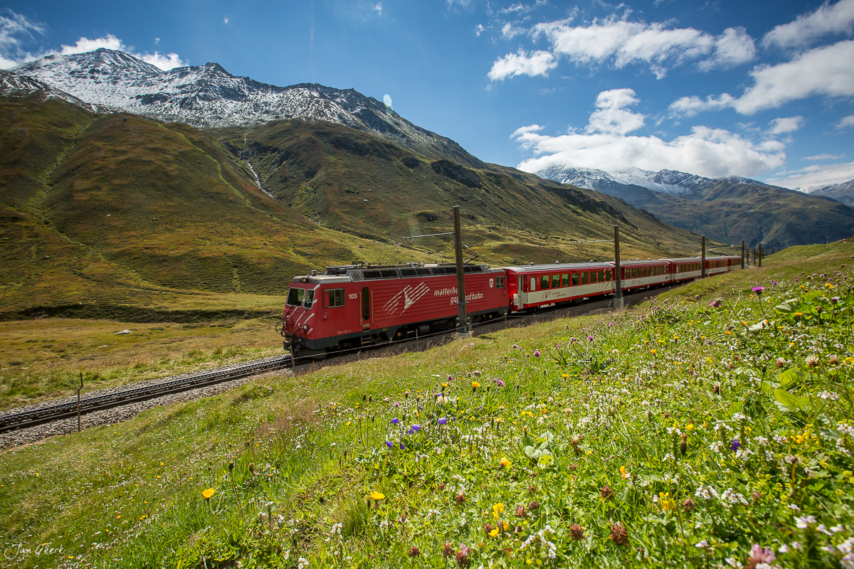 Roter Gotthard-Zug in Blütenwiese mit schneebedeckten Alpen, Frühjahr, Zentralschweiz – Jan Geerk Fotograf Basel