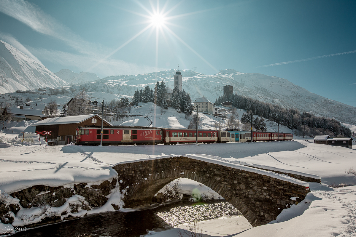 Roter Zug und Steinbrücke in winterlicher Berglandschaft, Hospental Oberalppass, sonniger Wintertag, Zentralschweiz Schweiz – Jan Geerk Fotograf Basel