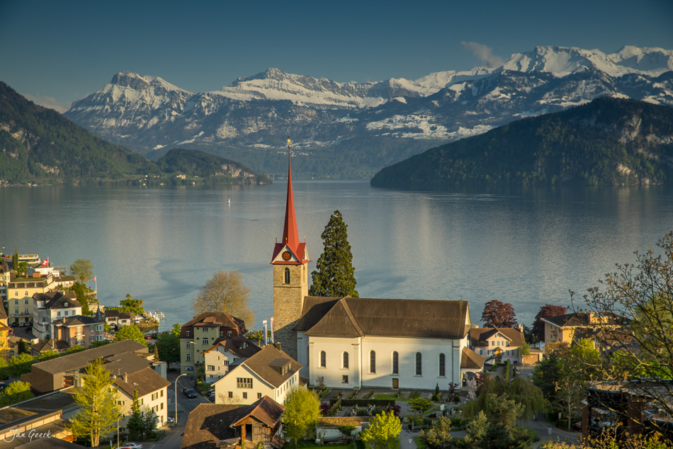 Dorfkirche mit rotem Dachspitz am Ufer des Vierwaldstättersee mit schneebedeckten Alpen im Hintergrund, Frühlingsstimmung, Zentralschweiz, Schweiz – Jan Geerk Fotograf Basel