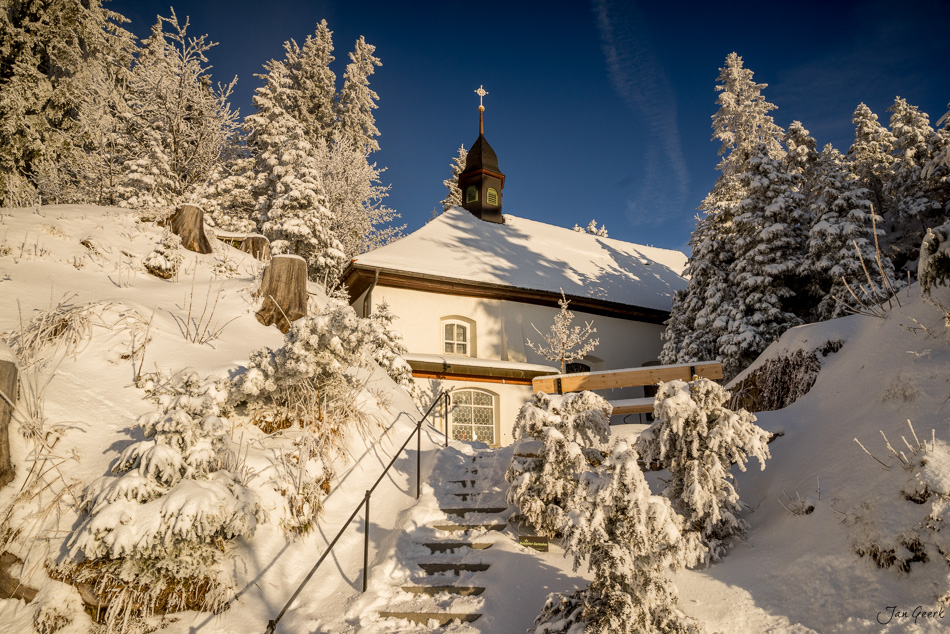 Schneebedeckte Kapelle mit Kreuz auf dem Dach umgeben von verschneiten Fichten auf der Rigi im Wintersonnenaufgang Zentralschweiz Schweiz – Jan Geerk Fotograf Basel