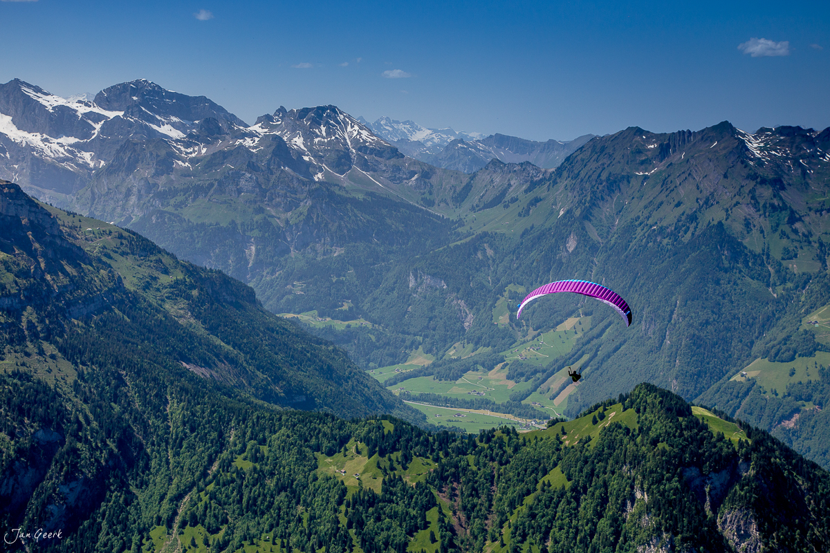Paraglider mit pink-violettem Schirm über der Seebodenalp in der Zentralschweiz, grüne Täler und schneebedeckte Berge bei Sonnenschein, Schweiz – Jan Geerk Fotograf Basel