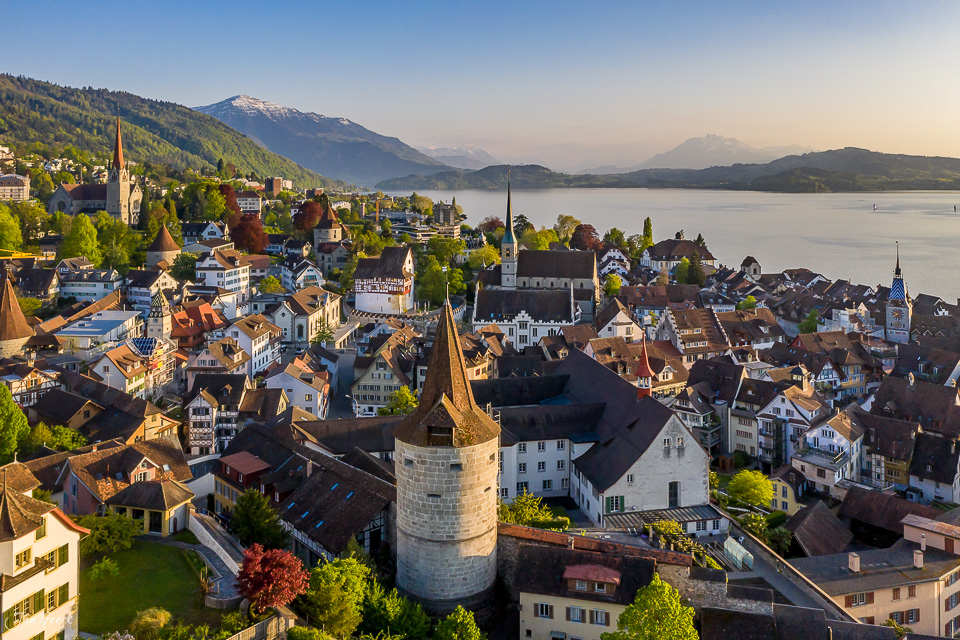 Luftaufnahme der Altstadt Zug mit Kirchtürmen, See und schneebedeckten Bergen im Frühling – Schweiz, Jan Geerk Fotograf Basel