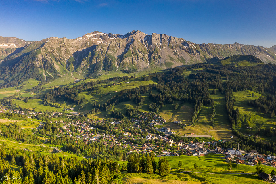 Luftaufnahme des Dorfes Sörenberg im Entlebuch mit grünen Wiesen, Wäldern und dramatischen Bergen im Hintergrund während sonnigen Sommertag, Schweiz – Jan Geerk Fotograf Basel