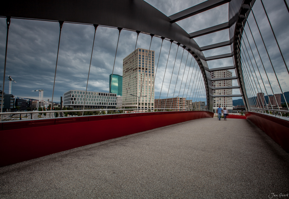 Moderne Fußgängerbrücke mit roten Geländern und Glasdach, Blick auf Hochhäuser und Skyline Zürich, bewölkter Himmel, Schweiz – Jan Geerk Fotograf Basel